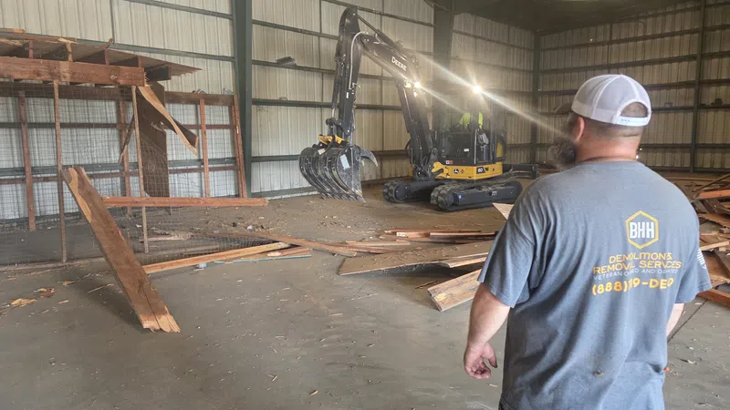 Worker in gray shirt overseeing indoor demolition with excavator clearing wooden debris inside metal building.