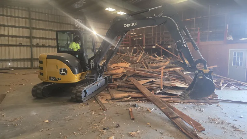 John Deere excavator inside warehouse clearing a large pile of wooden debris and lumber pieces scattered on the floor.