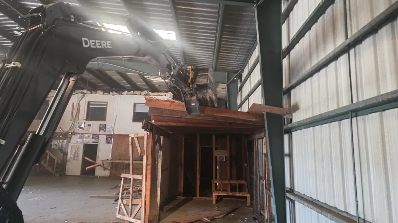 John Deere excavator demolishes wooden structure inside large metal warehouse building with debris on floor.