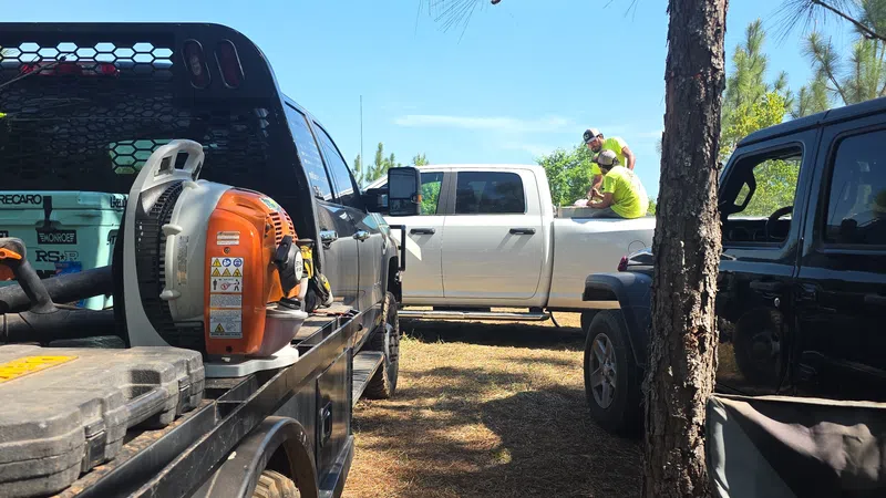 Work trucks parked on a dirt path with two workers in neon shirts sitting on a white pickup truck under a clear sky
