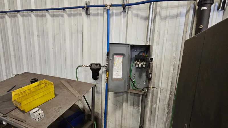 Industrial workspace with metal table, yellow toolbox, electrical panel, and blue piping on white wall.