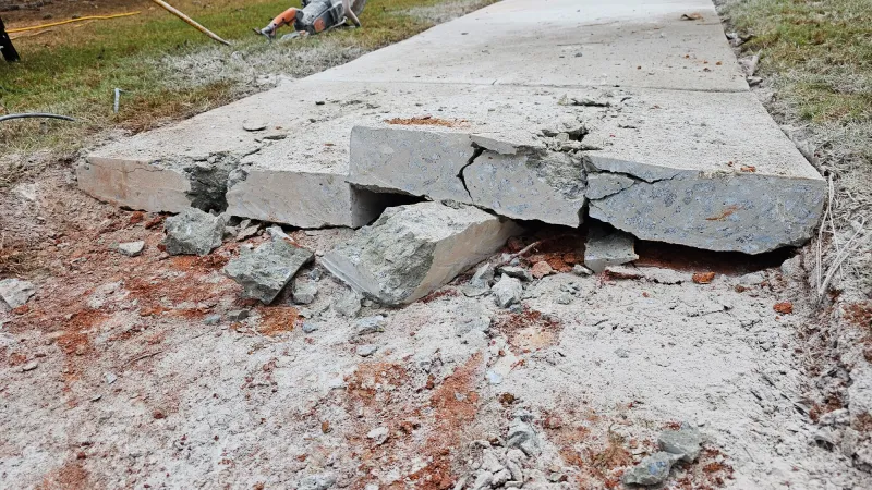 Cracked and broken concrete sidewalk with rubble and construction equipment nearby during repair work.