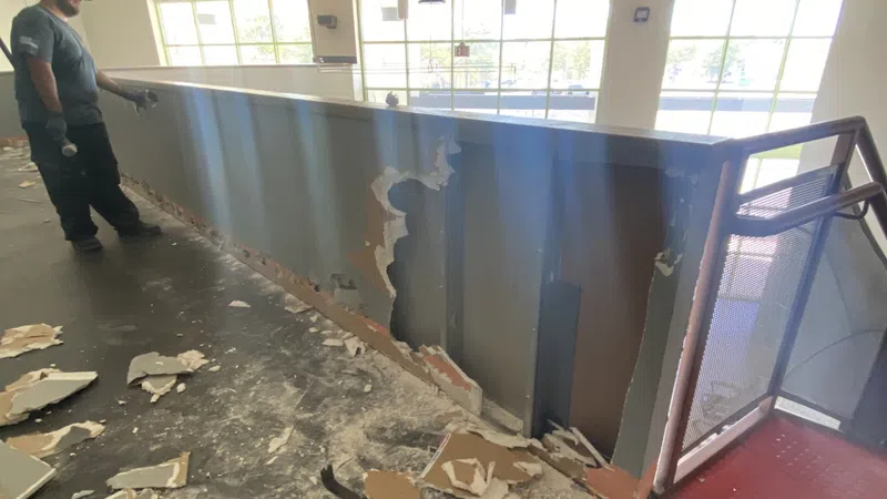 Worker standing next to damaged drywall railing with debris scattered on floor in a bright indoor space.