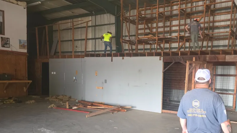 Three construction workers renovating a large indoor warehouse with wooden scaffolding and concrete floor under metal roof.