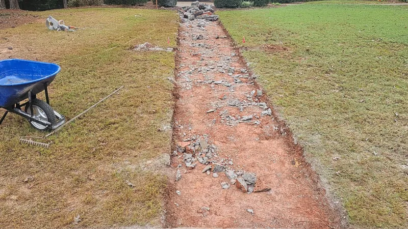 Excavated dirt path with scattered rocks and lawn tools, including a blue wheelbarrow and rake, on grass.
