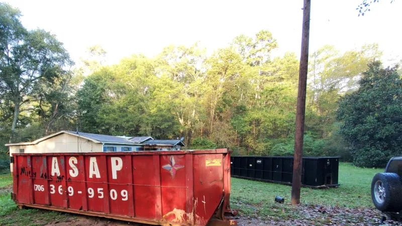 Red dumpster with ASAP phone number on grass near a house and trees in daytime light.