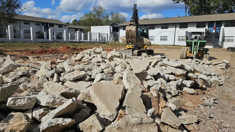 Construction site with broken concrete rubble in foreground and excavator and loader against residential buildings.