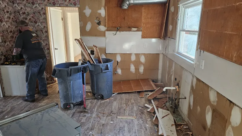 Room under renovation with exposed walls, debris on floor, two trash bins filled with wood, and a worker in the background
