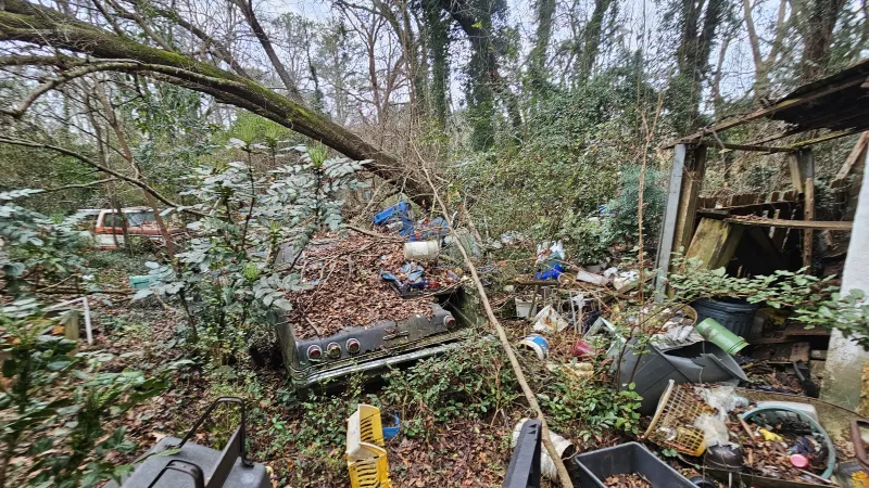 Overgrown forest area filled with scattered trash, old car parts, and abandoned household items near rundown structure.