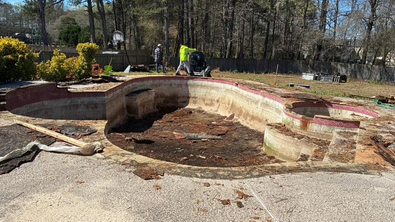 Empty, damaged backyard swimming pool undergoing cleanout and repair with workers and equipment nearby.