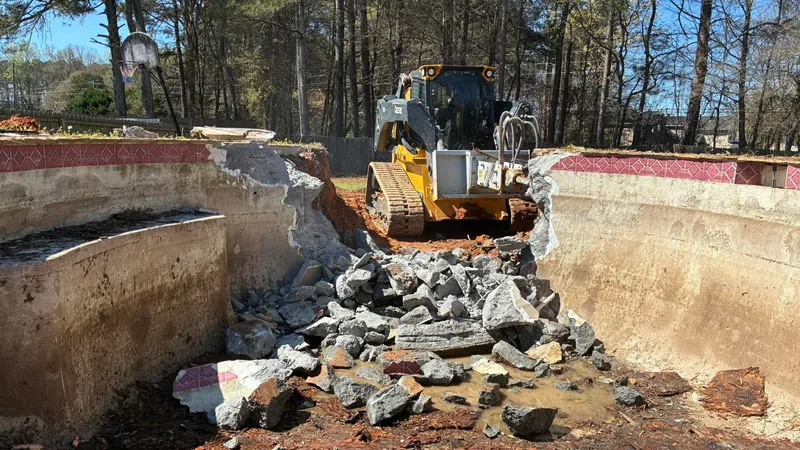 Bulldozer demolishing old concrete swimming pool with broken rubble inside on a sunny day.