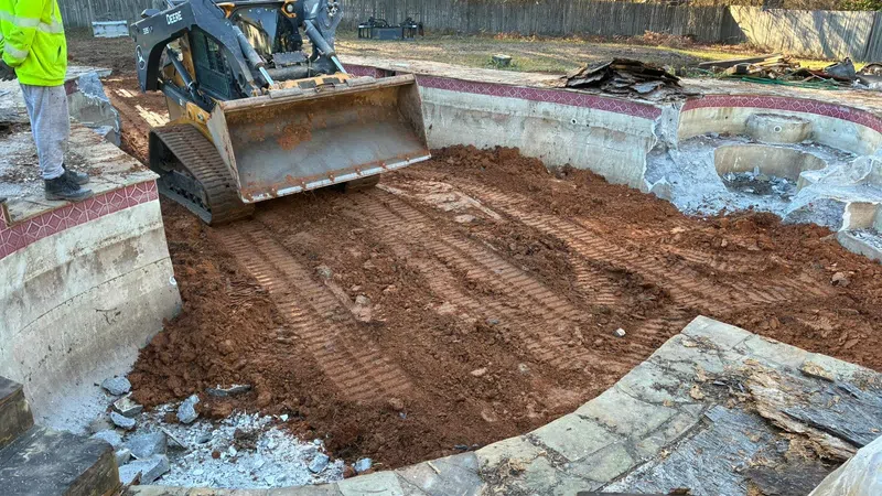Construction of an empty swimming pool with compacted dirt and a skid steer loader inside, worker observes the site.