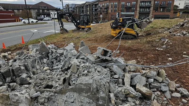 Construction site with demolished concrete debris, two yellow excavators, and residential buildings in background.