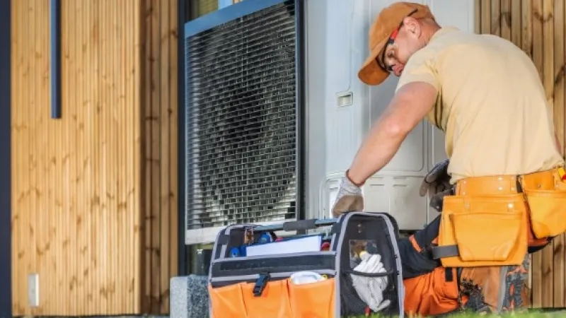 Technician in orange cap and gloves repairing outdoor HVAC unit next to wooden house wall with open tool bag