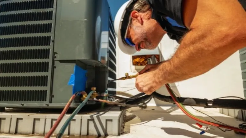 Technician wearing hard hat repairing outdoor air conditioning unit under clear blue sky.