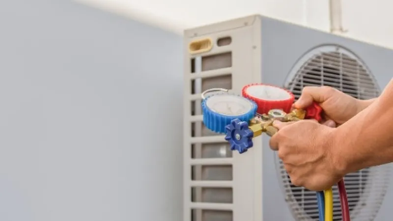 Technician holding HVAC gauges while servicing an air conditioning unit outdoors on a sunny day