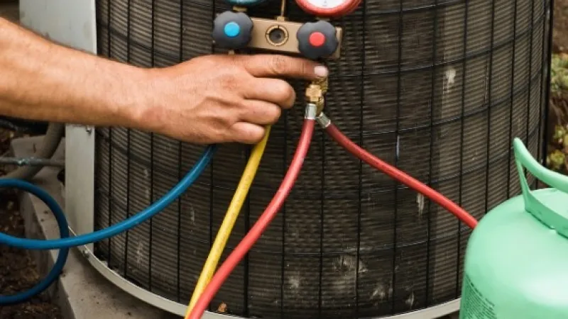 Technician checking HVAC air conditioning unit pressure with manifold gauge and refrigerant tank outdoors.