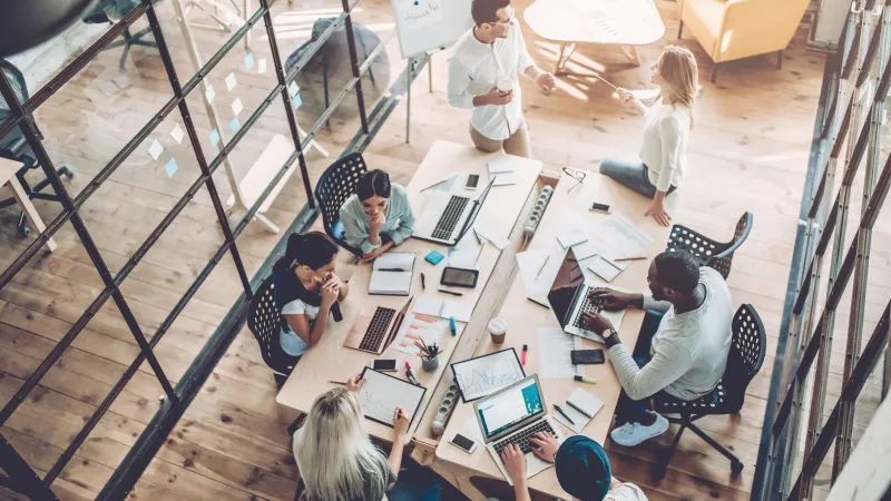 Top view of diverse team collaborating around a table with laptops, notebooks, and charts in a modern office space.