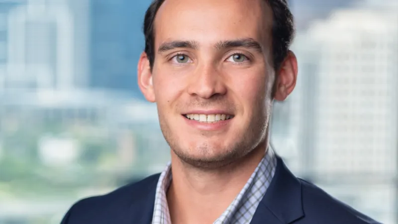 Smiling young man in a navy blazer and checked shirt with a blurred cityscape background.