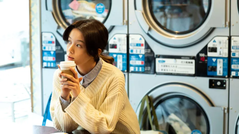 Woman in cozy sweater enjoying coffee while waiting at a laundromat with washing machines in the background