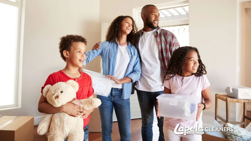Happy family in casual clothes smiling and holding moving boxes and a teddy bear in a bright new home.