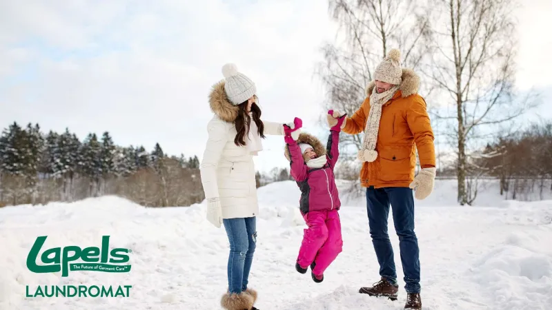 Family of three in winter clothes playing outdoors in snowy landscape with trees in background.