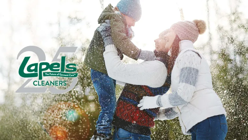 A family enjoys a snowy day outdoors with parents lifting their child, with Lapels Cleaners 25th anniversary logo.