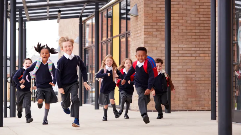 Group of diverse schoolchildren in uniforms running happily outside a modern school building on a sunny day