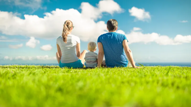 Family of three sitting on grass facing blue sky with white clouds on a sunny day