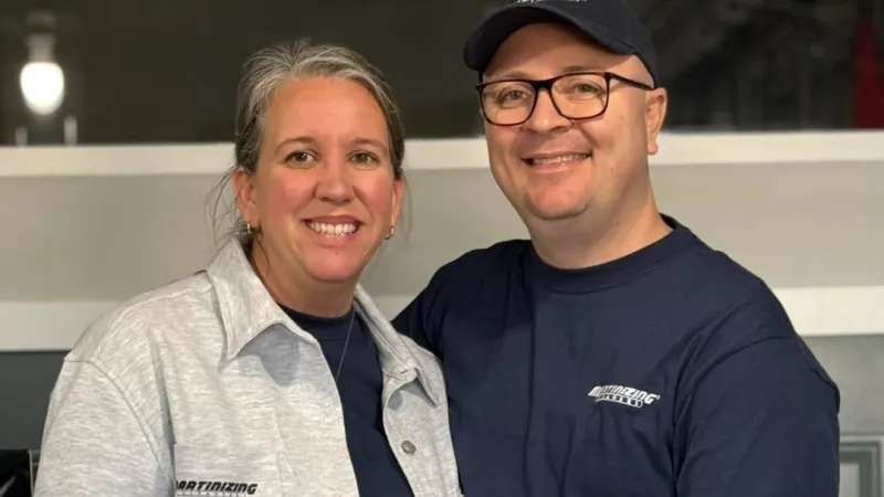 Smiling couple wearing matching casual shirts and glasses posing indoors, showcasing company logo apparel.