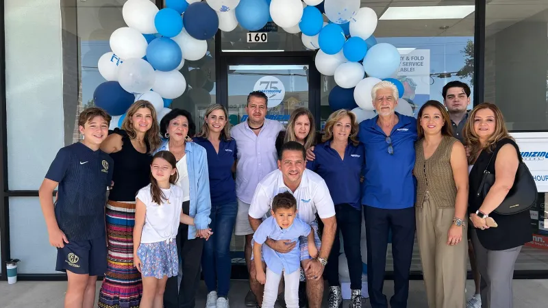 Group of diverse people smiling under a blue and white balloon arch outside a storefront on a sunny day