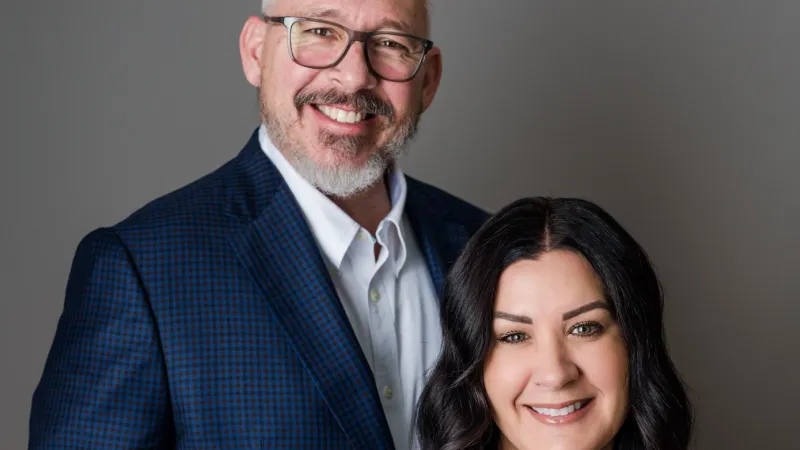 Smiling professional man in blue blazer and woman in white blouse posing against gray background.