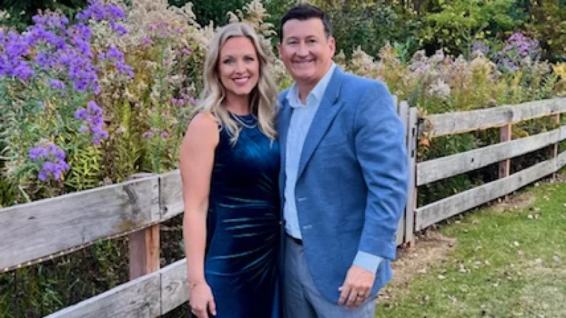 A smiling couple dressed formally standing beside a wooden fence with colorful flowers and trees in the background.