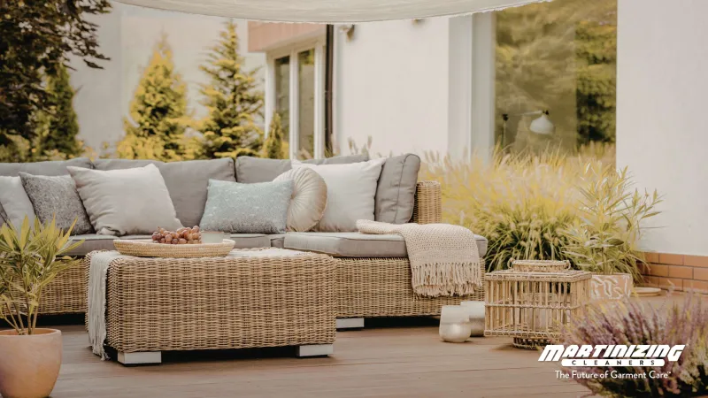 Outdoor wicker patio sofa with cushions and throw blanket on wooden deck surrounded by plants and lanterns.