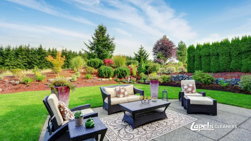 Outdoor patio with wicker furniture, cushions, coffee table, garden plants, and blue sky in a landscaped backyard.