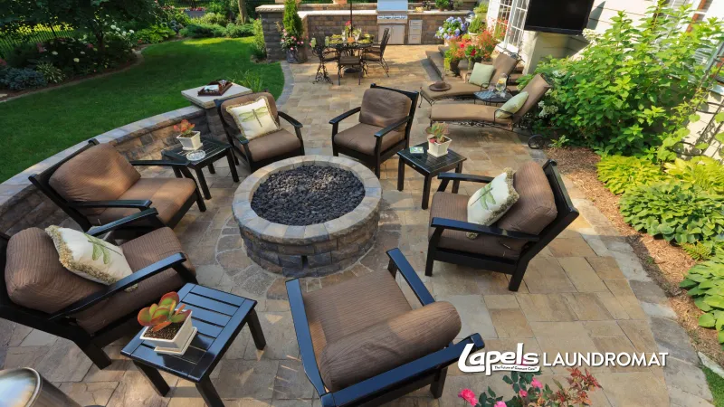 Outdoor patio with cushioned chairs around a stone fire pit and a garden seating area in the background