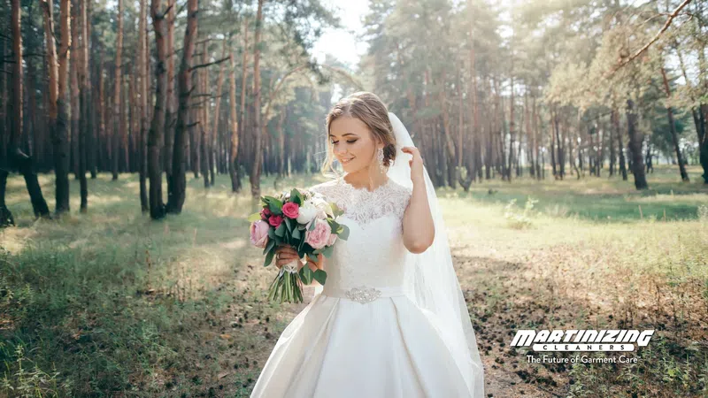 Bride in white wedding dress holding bouquet in sunlit forest clearing, smiling and looking down.