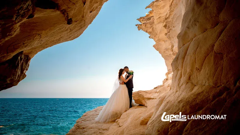 Bride and groom standing on rocky cliff by the ocean, embracing on a sunny day during their wedding.