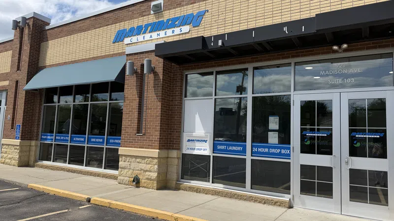 Exterior view of Martinizing Cleaners storefront with signage, blue awning, and 24-hour drop box on a sunny day.