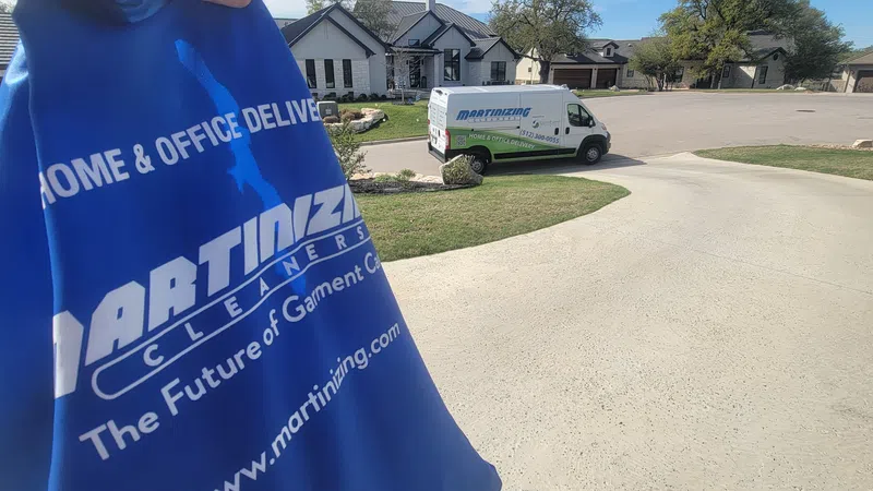 Hand holding blue garment bag from Martinizing Cleaners with delivery van and suburban home in background.