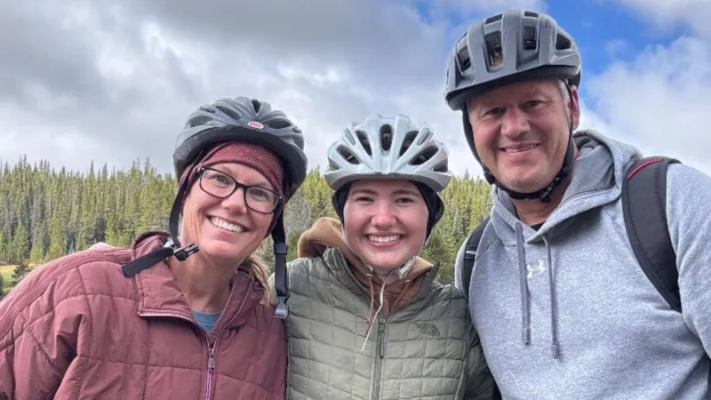 Three smiling people wearing bike helmets pose outdoors with pine trees and cloudy sky in the background