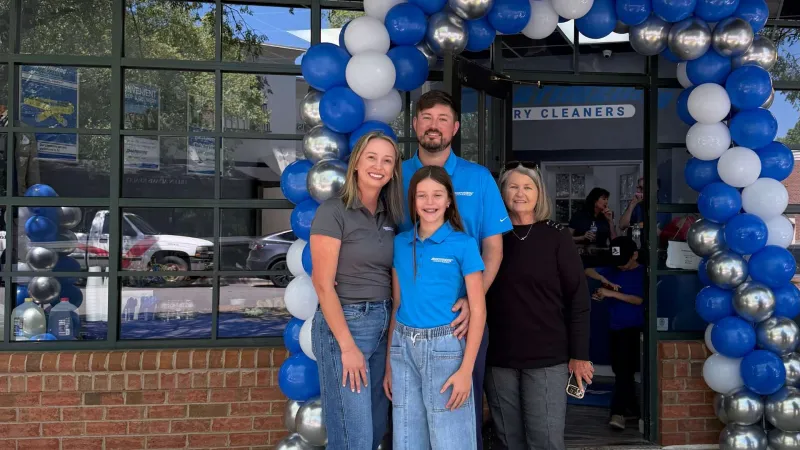 Four people posing under a blue, white, and silver balloon arch in front of a dry cleaners store.
