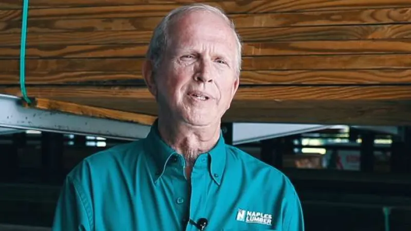 Man wearing teal Naples Lumber uniform shirt standing in front of stacked wooden boards in a warehouse.