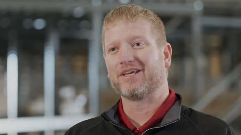 Smiling man with red hair and beard wearing black jacket indoors with blurred industrial background
