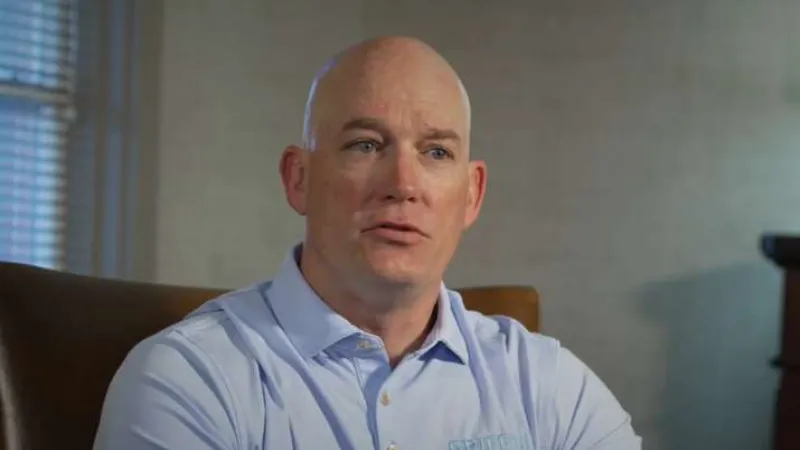 Bald man in light blue collared shirt sitting indoors with serious expression, neutral background and window blinds.