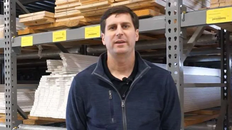 Man in blue jacket standing in front of shelves with stacked wooden planks and boards in a warehouse.
