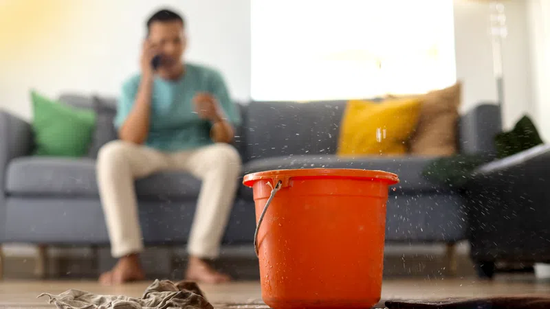 A bright orange bucket collects water on a wooden floor while a person talks on the phone in the background.