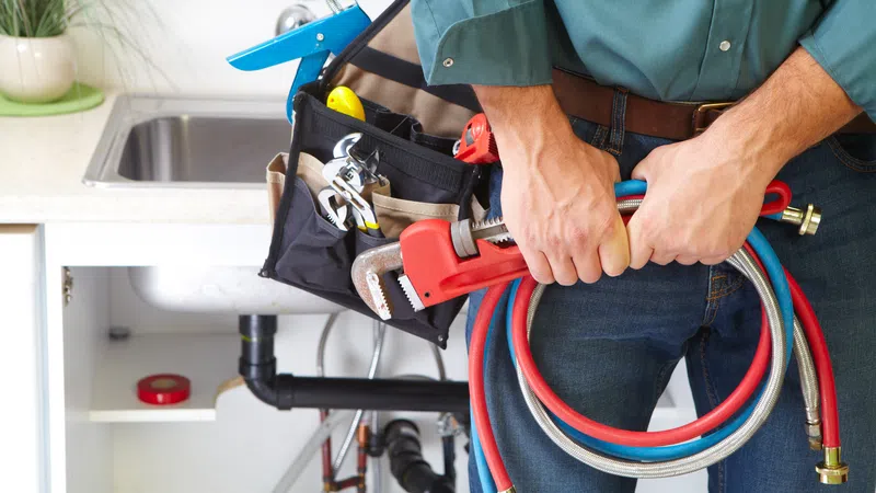 Plumber holding wrench and coiled hoses standing next to open kitchen sink with visible pipes and tool belt.