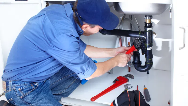 Plumber in blue uniform fixing kitchen sink pipes with tools and toolbox under cabinet.