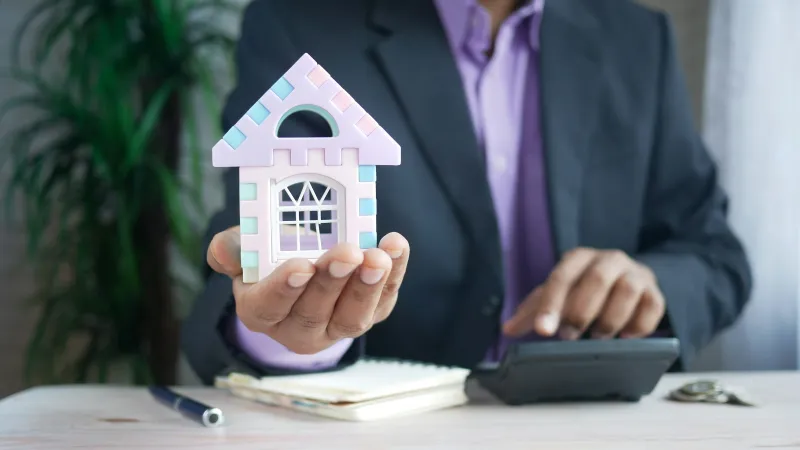 Businessman holding a small pastel-colored house model with notebook, pen, and calculator on desk.